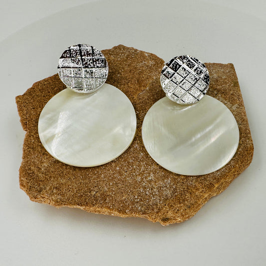 Pair of earrings with white round stones and silver checkered tops on a brown rock.